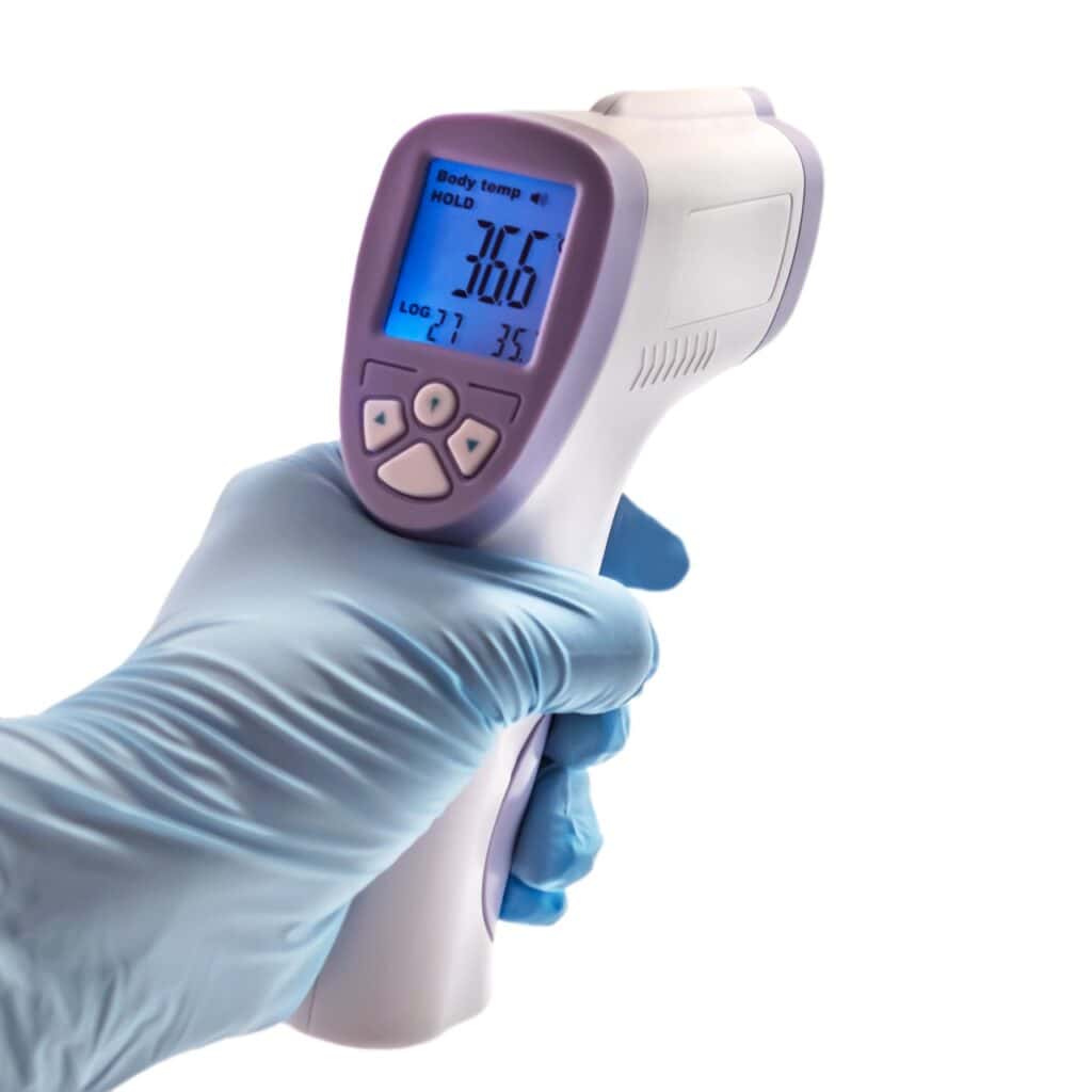 Mano con guante azul sosteniendo termómetro infrarrojo sin contacto para medición de temperatura corporal, mostrando lectura de 36.6°C
