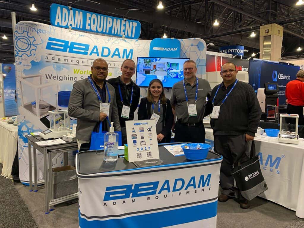 Equipo de Adam Equipment posando en stand de feria Pittcon con balanzas, catálogo 2023 y material promocional