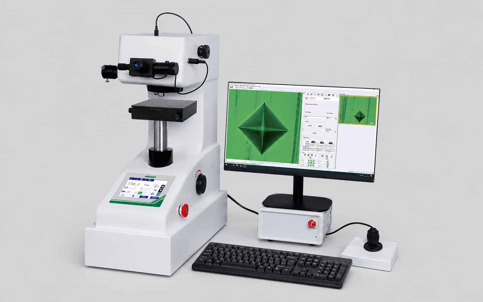 Durómetro digital con sistema de medición automatizado, pantalla de control y monitor externo mostrando la huella de ensayo, sobre fondo gris claro homogéneo en entorno de laboratorio.